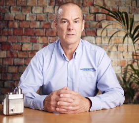 Andrew Goode sat at a wooden desk with a hydrogen pressure regulator in front of him, and a brick wall and green plant behind him.