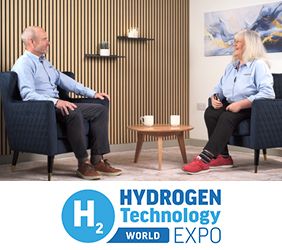 Susie Muir and Andrew Goode sat in blue tub chairs having a discussion with wood panneling on the wall behind them and a small round wooden coffee table in between them with two white tea cups sat on top.